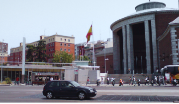 Aparcamiento en Moncloa Madrid 2026: aparcar cerca del intercambiador. La foto muestra una calle con en primer plano un coche que anda y el fondo es el complejo del intercambiador de Moncloa en Madrid.