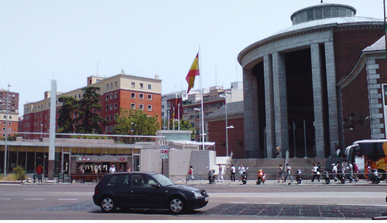 Aparcamiento en Moncloa Madrid 2026: aparcar cerca del intercambiador. La foto muestra una calle con en primer plano un coche que anda y el fondo es el complejo del intercambiador de Moncloa en Madrid.